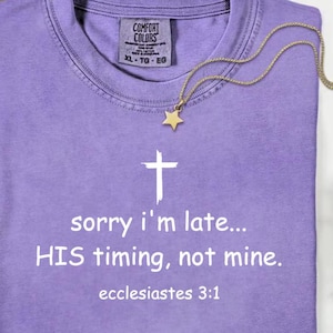 May include: A purple t-shirt with the text "sorry i'm late... HIS timing, not mine. ecclesiastes 3:1" and a white cross graphic. A gold star pendant necklace is draped across the shirt. The shirt has a Comfort Colors tag.