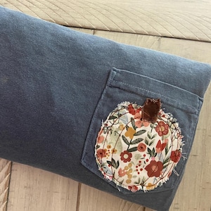 May include: A blue rectangular pillow with a pocket featuring a floral pumpkin applique. The pumpkin design includes red, orange, and yellow flowers with green leaves. The stem is a small piece of brown fabric.
