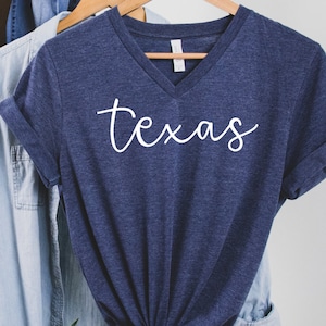 K&ouml;nnte beinhalten: Ein marineblaues V-Ausschnitt-T-Shirt mit dem Wort "texas" in wei&szlig;er Schreibschrift. Das Shirt h&auml;ngt an einem Holzb&uuml;gel mit anderen Kleidungsst&uuml;cken im Hintergrund.