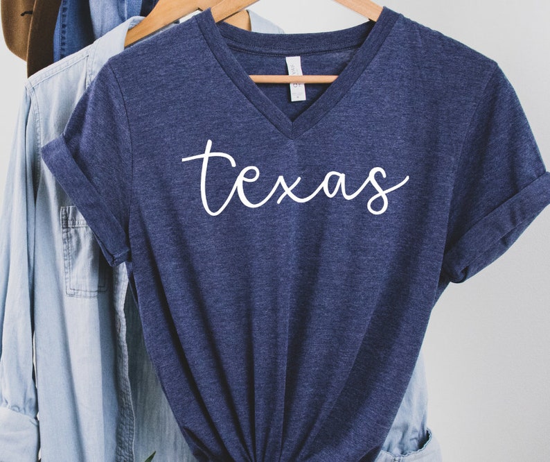 K&ouml;nnte beinhalten: Ein marineblaues V-Ausschnitt-T-Shirt mit dem Wort "texas" in wei&szlig;er Schreibschrift. Das Shirt h&auml;ngt an einem Holzb&uuml;gel mit anderen Kleidungsst&uuml;cken im Hintergrund.