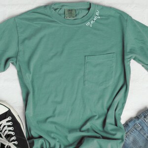 K&ouml;nnte beinhalten: Ein blaues T-Shirt mit einem Taschenfach und dem Wort "mama" auf der Vorderseite gestickt.