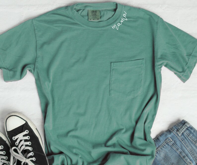 K&ouml;nnte beinhalten: Ein blaues T-Shirt mit einem Taschenfach und dem Wort "mama" auf der Vorderseite gestickt.