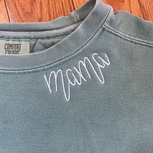 K&ouml;nnte beinhalten: Ein hellblauer Pullover mit dem Wort "mamma" in wei&szlig; auf der Vorderseite gestickt.