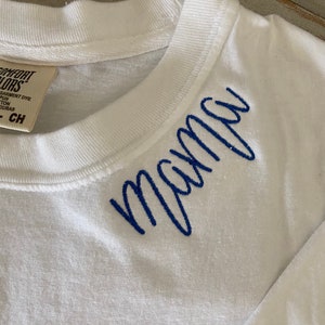 K&ouml;nnte beinhalten: Wei&szlig;es Langarm-T-Shirt mit blau gesticktem Text "mama" am Ausschnitt.