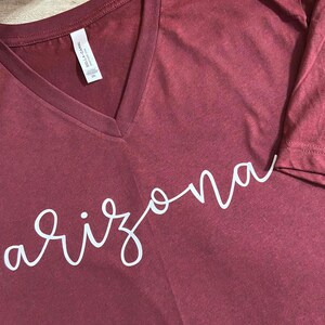 K&ouml;nnte beinhalten: Burgunderfarbenes V-Ausschnitt-T-Shirt mit dem Wort "Arizona" in wei&szlig;er Schrift. Das Shirt ist aus einem weichen Material gefertigt und hat ein Etikett am inneren Kragen. Das Shirt hat die Gr&ouml;&szlig;e 2XL.