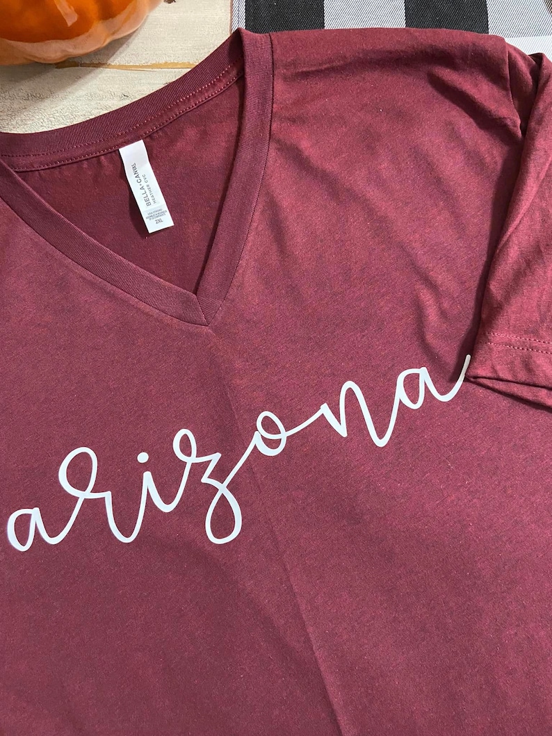 K&ouml;nnte beinhalten: Burgunderfarbenes V-Ausschnitt-T-Shirt mit dem Wort "Arizona" in wei&szlig;er Schrift. Das Shirt ist aus einem weichen Material gefertigt und hat ein Etikett am inneren Kragen. Das Shirt hat die Gr&ouml;&szlig;e 2XL.