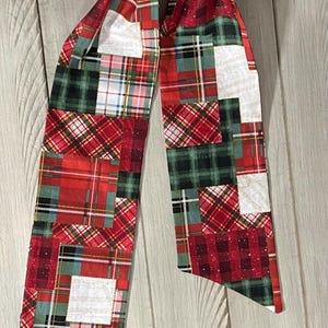 May include: A decorative scarf featuring a patchwork design with red, green, and white plaid patterns. The scarf is made of fabric and has a long, slender shape, perfect for accessorizing. The scarf is shown against a light wood background.