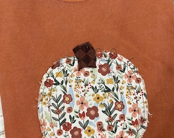 May include: Rust-colored sweatshirt with a floral pumpkin applique. The pumpkin is filled with a variety of flowers in shades of red, yellow, and brown. The sweatshirt has a crew neck and the brand "Comfort Colors" is visible.