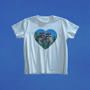 May include: A white short-sleeve t-shirt featuring a heart-shaped graphic on the chest. The graphic depicts two raccoons surrounded by green foliage against a blue background. The text Marigai Maria is printed in small black letters on the inner collar.