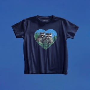 May include: A navy blue short-sleeve t-shirt featuring a central heart-shaped graphic. The graphic depicts two raccoons nestled within green foliage. The interior neckline displays the text Marigold Maria in white lettering on the dark fabric.