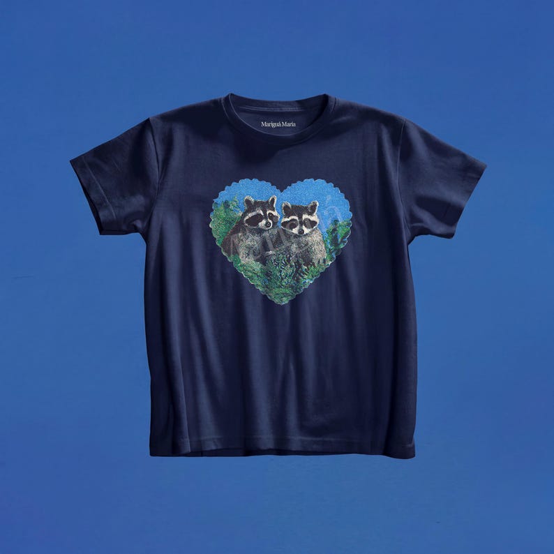 May include: A navy blue short-sleeve t-shirt featuring a central heart-shaped graphic. The graphic depicts two raccoons nestled within green foliage. The interior neckline displays the text Marigold Maria in white lettering on the dark fabric.