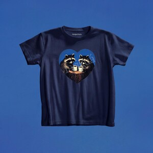 May include: A navy blue short-sleeve t-shirt featuring a central graphic of two raccoons sitting at a tree stump, each holding a white mug. The graphic is enclosed within a blue heart shape. The brand name Marig&uacute; Maria is printed on the inner neck.