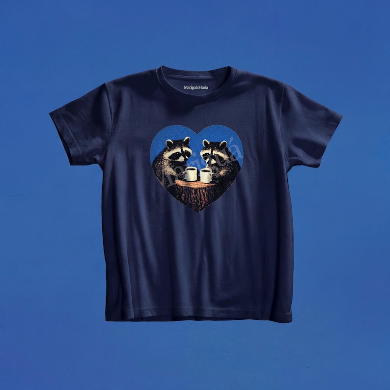 May include: A navy blue short-sleeve t-shirt featuring a central graphic of two raccoons sitting at a tree stump, each holding a white mug. The graphic is enclosed within a blue heart shape. The brand name Marig&uacute; Maria is printed on the inner neck.