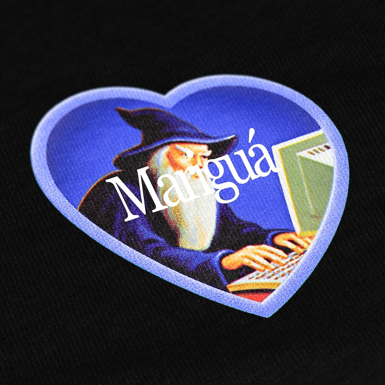 May include: A black fabric surface featuring a heart-shaped graphic print. The design depicts a wizard with a long white beard wearing a dark blue robe, typing on a computer keyboard. The white text "Marigu&aacute;" is printed across the center.