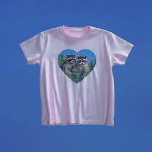 May include: A light pink short-sleeve t-shirt featuring a central graphic of two raccoons nestled within a blue heart-shaped frame. The raccoons are depicted in shades of gray, black, and white, surrounded by green foliage inside the heart design.