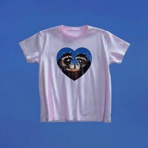 May include: A light pink short-sleeve t-shirt featuring a central blue heart graphic. Inside the heart, two raccoons are depicted sitting on a tree stump, each holding a small white mug. The design is centered on the chest of the garment.