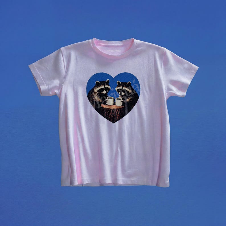 May include: A light pink short-sleeve t-shirt featuring a central blue heart graphic. Inside the heart, two raccoons are depicted sitting on a tree stump, each holding a small white mug. The design is centered on the chest of the garment.