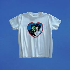 Może przedstawiać: Biały t-shirt z krótkim rękawem z centralną grafiką w kształcie serca. Wewnątrz czerwonego obramowania serca znajduje się ilustracja potwora o zielonej skórze i kobiety w białej sukni. Napis Marigai Maria jest nadrukowany na wewnętrznej stronie dekoltu.