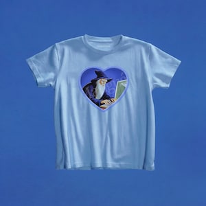May include: A light blue short-sleeve t-shirt featuring a central heart-shaped graphic. The graphic depicts a wizard with a long white beard and a dark blue hat typing on a computer keyboard. The design is set against a dark blue background.