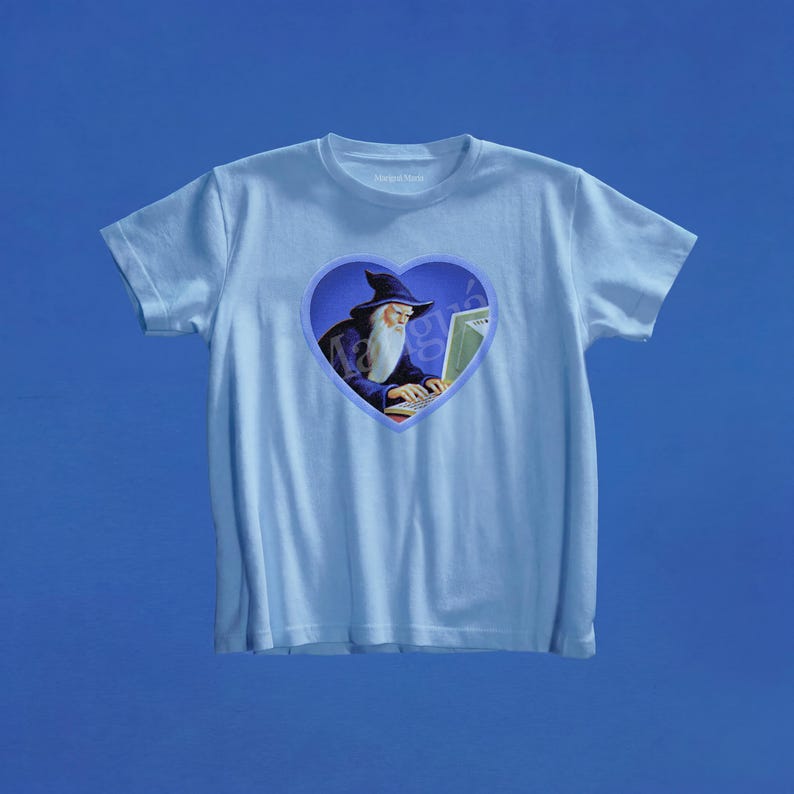 May include: A light blue short-sleeve t-shirt featuring a central heart-shaped graphic. The graphic depicts a wizard with a long white beard and a dark blue hat typing on a computer keyboard. The design is set against a dark blue background.