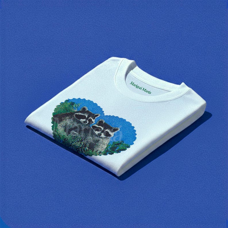 May include: A white crewneck sweatshirt featuring a graphic of two raccoons nestled within a blue heart-shaped background surrounded by green foliage. The inner collar area displays the green text Marigui Maria printed on the fabric.