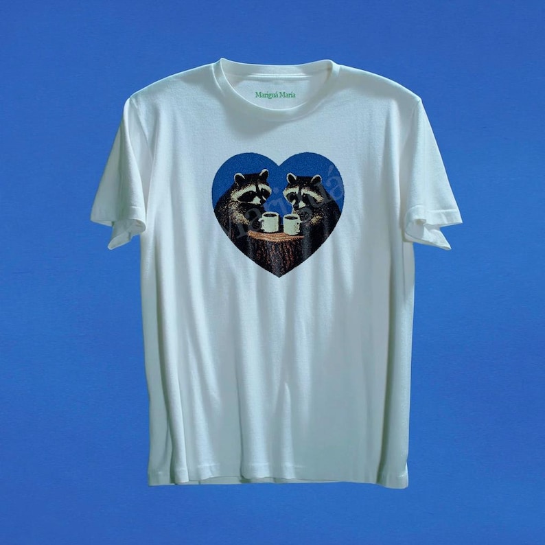 May include: A white short-sleeved t-shirt featuring a blue heart-shaped graphic on the chest. Inside the heart, two raccoons are depicted sitting on a tree stump, each holding a white mug. The text Marigui Maria is printed inside the neck collar.