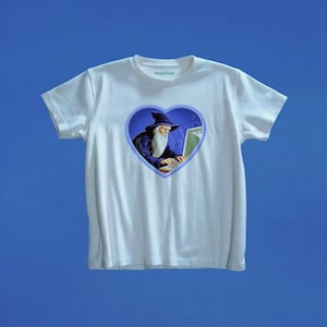 Op de afbeelding: Een wit T-shirt met korte mouwen en een centraal hartvormig grafisch ontwerp. Binnenin het hart typt een tovenaar met een lange witte baard en een donkerblauwe hoed op een computer. De tekst Marigai Maria staat in kleine groene letters.