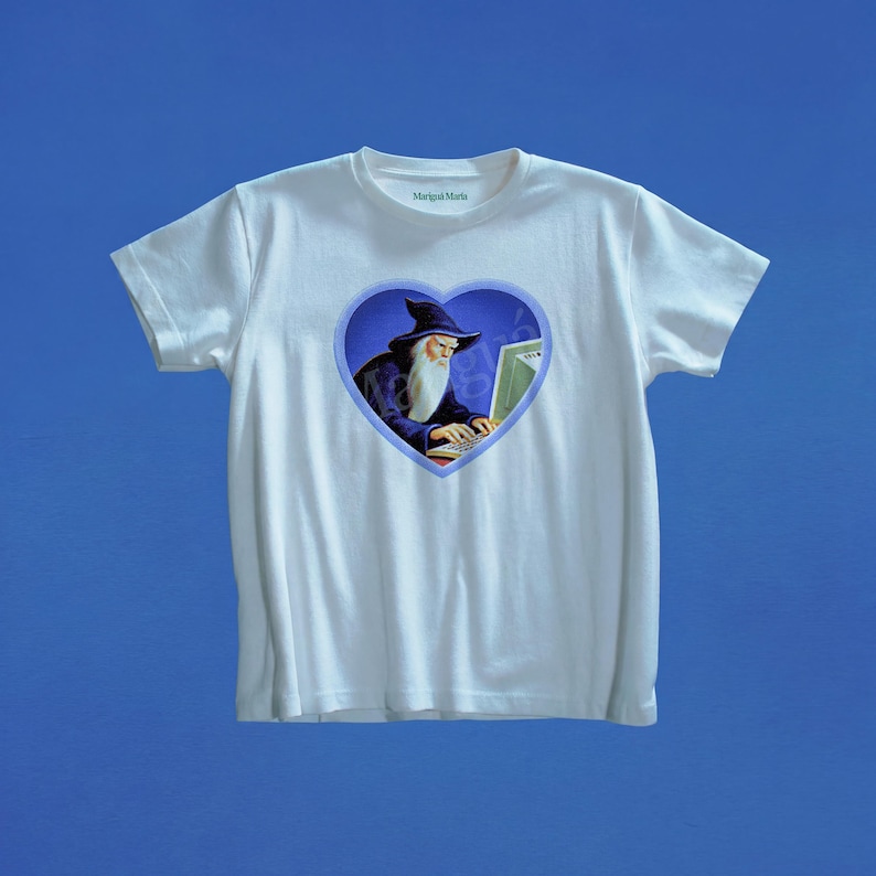 May include: A white short-sleeved t-shirt featuring a central heart-shaped graphic. Inside the heart, a wizard with a long white beard and a dark blue hat is typing on a computer. The text Marigai Maria is printed in small green letters at the neckline.