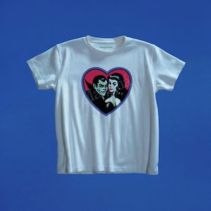 May include: A white short-sleeved t-shirt featuring a central heart-shaped graphic. Inside the heart is a vintage-style illustration of a vampire and a woman with dark hair. The text Maripai Maria is printed on the inner neckline of the garment.