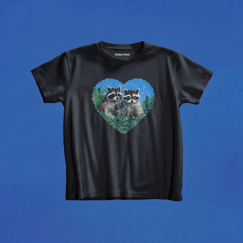 May include: A black short-sleeved t-shirt featuring a heart-shaped graphic on the chest. The graphic depicts two raccoons nestled within green foliage against a blue background. The text Marigul Maria is printed in white on the inside neck label.