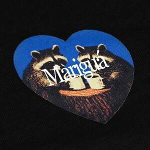 May include: A black fabric surface featuring a heart-shaped graphic print. The design displays two raccoons in dark brown and black tones, holding white mugs against a blue background. The white text Marigu&aacute; is printed across the center.