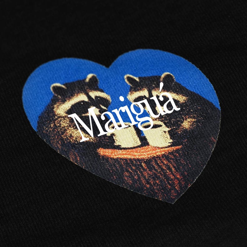 May include: A black fabric surface featuring a heart-shaped graphic print. The design displays two raccoons in dark brown and black tones, holding white mugs against a blue background. The white text Marigu&aacute; is printed across the center.