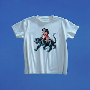 Może przedstawiać: Biały t-shirt z krótkim rękawem z nadrukiem graficznym przedstawiającym osobę z długimi ciemnymi włosami w czerwonym body, jadącą na czarnej panterze w niebieskie cętki. Na wewnętrznej stronie kołnierzyka widnieje napis Marigai Maria małymi czarnymi literami.