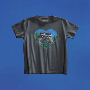 May include: A charcoal gray short-sleeve t-shirt featuring a heart-shaped graphic on the chest. The graphic depicts two raccoons nestled within green foliage against a blue background. The brand name Marigai Maria is printed in white inside the neck.