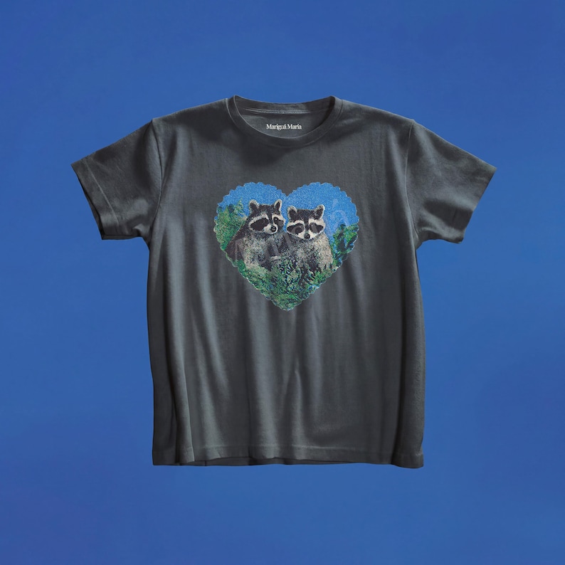 May include: A charcoal gray short-sleeve t-shirt featuring a heart-shaped graphic on the chest. The graphic depicts two raccoons nestled within green foliage against a blue background. The brand name Marigai Maria is printed in white inside the neck.
