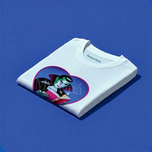 May include: A white crew neck t-shirt featuring a graphic of a vampire with green skin reading a book inside a pink heart-shaped frame. The text Marigui Maria is printed in green on the inside collar area of the folded garment.