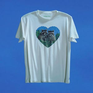 May include: A white short-sleeved t-shirt featuring a central heart-shaped graphic containing two raccoons surrounded by green foliage against a blue background. The inner collar displays the green text Marigui Maria in a simple serif font.