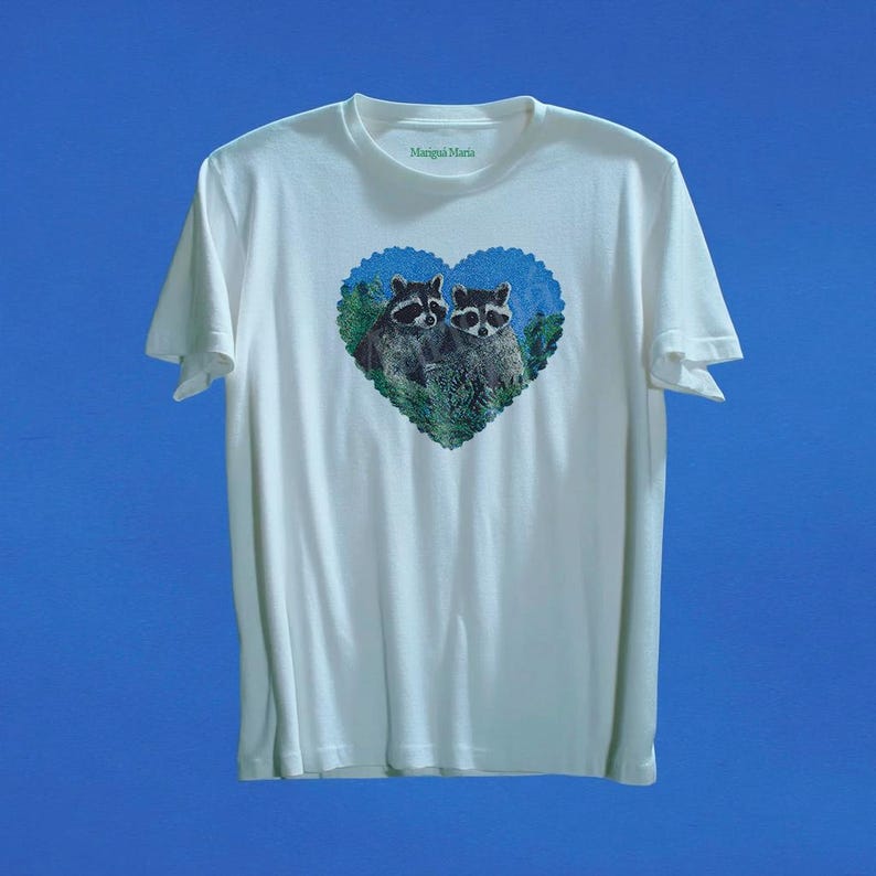 May include: A white short-sleeved t-shirt featuring a central heart-shaped graphic containing two raccoons surrounded by green foliage against a blue background. The inner collar displays the green text Marigui Maria in a simple serif font.