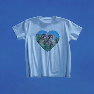 May include: A light blue short-sleeve t-shirt featuring a central graphic of two raccoons nestled within a heart-shaped frame. The heart design includes green foliage and a blue background, with the raccoons depicted in their natural grey and black colors.