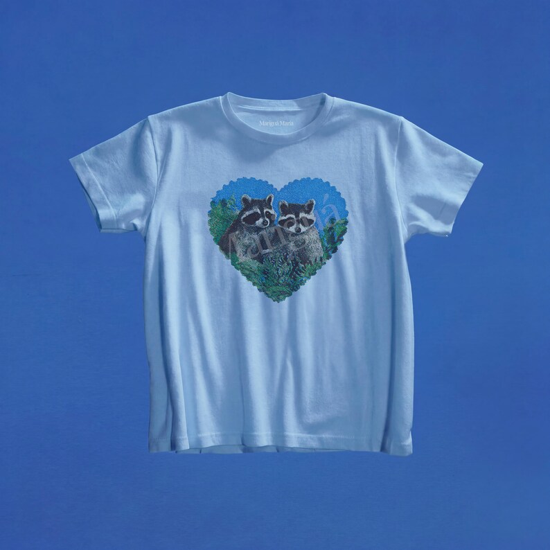 May include: A light blue short-sleeve t-shirt featuring a central graphic of two raccoons nestled within a heart-shaped frame. The heart design includes green foliage and a blue background, with the raccoons depicted in their natural grey and black colors.