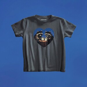May include: A dark gray short-sleeve t-shirt featuring a central heart-shaped graphic. The design depicts two raccoons sitting on a tree stump, each holding a white mug. The text Marigai Maria is printed in white on the inside of the shirt collar.