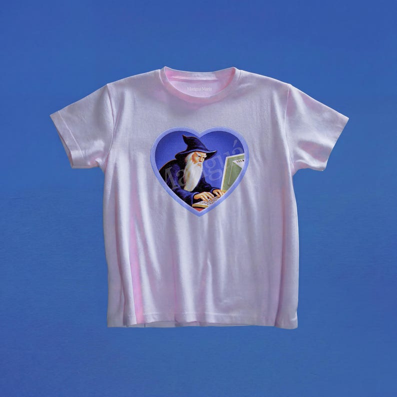 May include: A light pink short-sleeve t-shirt featuring a central heart-shaped graphic. Inside the heart, a wizard with a long white beard and a dark blue pointed hat is depicted typing on a computer keyboard against a dark blue background.