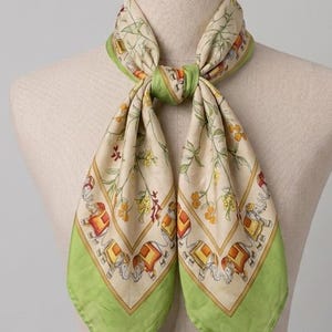 May include: A square scarf tied around a mannequin neck, featuring a cream-coloured centre with a repeating pattern of elephants and floral vines, bordered by a solid light green frame with a thin dark green line along the outer edge.