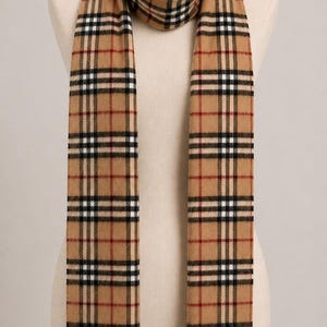 May include: A tan scarf featuring a classic plaid pattern in black, white, and red stripes. The scarf has fringed ends and measures 147 x 30 cm. It is draped around a mannequin neck, displaying the rectangular shape and the repeating check design.