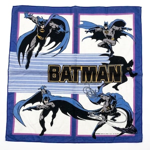 May include: A square blue and white fabric bandana featuring four distinct illustrations of Batman in action poses. The centre displays the word BATMAN in bold yellow letters with a black outline, accompanied by the text TM & © 1989 DC Comics Inc.