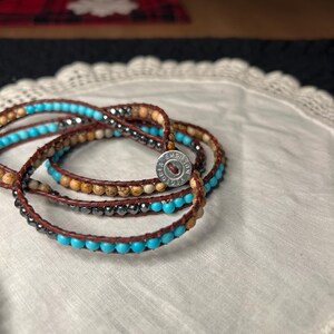 May include: Wrap bracelets with turquoise, brown, and gray beads. The bracelets are made of leather and have a metal clasp. The bracelets are displayed on a white lace doily.