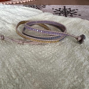 May include: A wrap bracelet with three strands of beads in gold, grey, and lavender. The bracelet is wrapped around itself and has a button closure. The bracelet is on a light green textured surface.