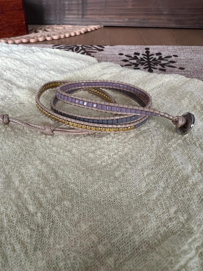 May include: A wrap bracelet with three strands of beads in gold, grey, and lavender. The bracelet is wrapped around itself and has a button closure. The bracelet is on a light green textured surface.