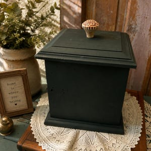 May include: A square wooden box painted in a matte dark charcoal finish with a flat lid and a decorative, light-colored, textured knob. The box features a stepped edge design on the lid and a solid base, presenting a clean, geometric structure.