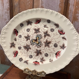May include: An oval cream-colored ceramic platter with a scalloped edge. The center features a detailed botanical pattern of dark brown leaves, red flowers, and black berries arranged in a symmetrical design radiating from a central floral motif.